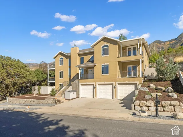Stucco exterior featuring multiple balconies with black railings, a three-car garage, and a tiered rock retaining wall