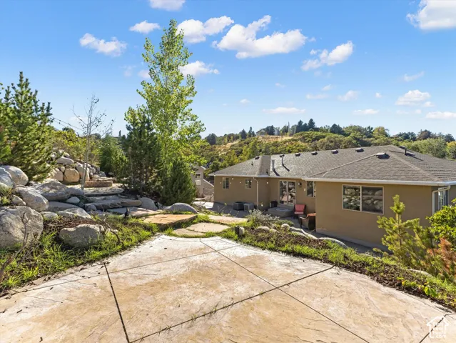 Stucco exterior with multiple windows, leading to an extensive stamped concrete patio, rock landscaping, and mature trees