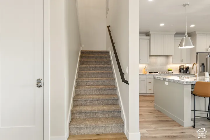 Carpeted staircase with dark handrail, white interior door with contemporary hardware, wood-finish flooring, and a kitchen featuring light cabinetry and a stone island countertop