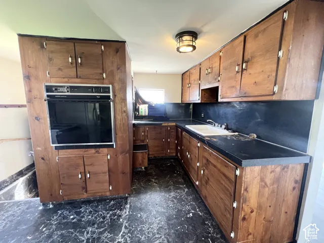 Built-in wall oven, wood-finish cabinetry, dark countertops, and a single-basin sink