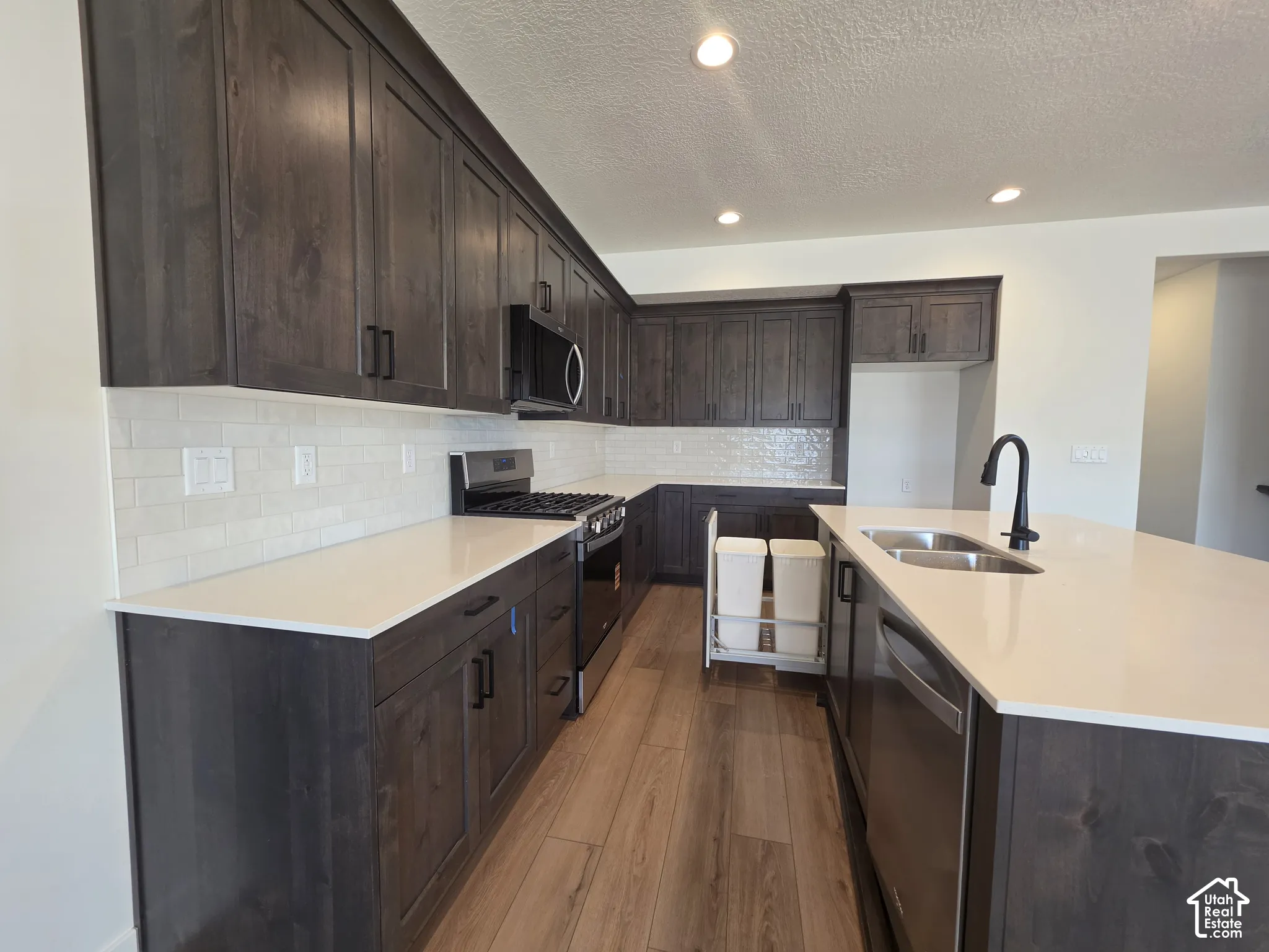 Kitchen featuring wood-finish flooring, dark wood cabinetry, white countertops, a subway tile backsplash, and a stainless steel sink with a matte black faucet