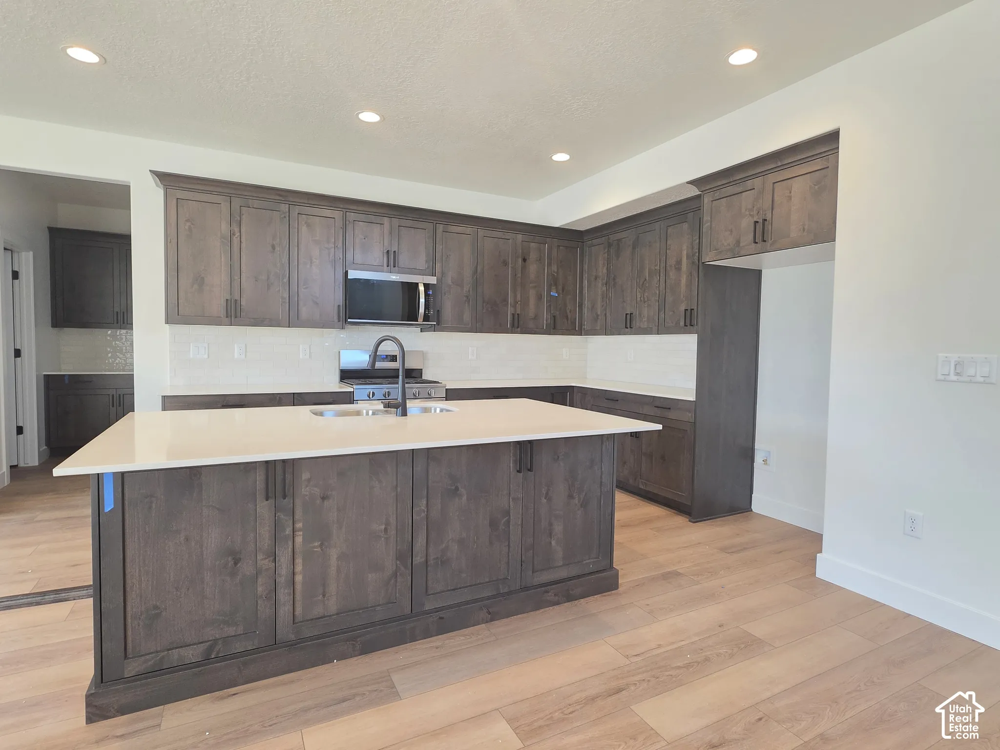 Spacious kitchen featuring an expansive center island, extensive dark wood-finish cabinetry, stainless steel appliances, a subway tile backsplash, and light wood-finish flooring