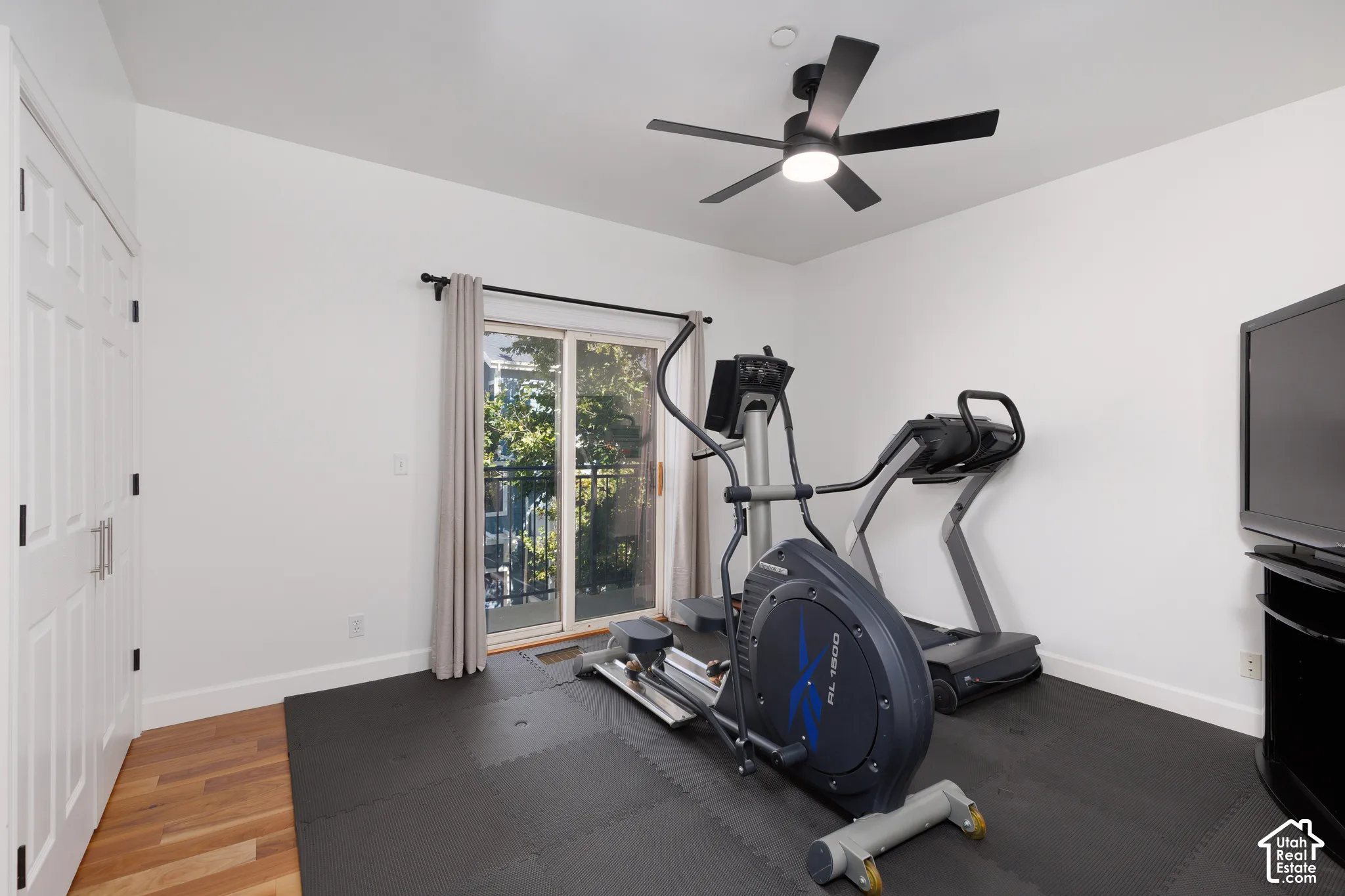 White walls with natural wood-finish flooring, ceiling fan with integrated lighting, and a sliding glass door to an outdoor balcony