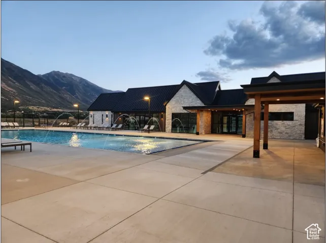 Resort-style pool with water features, stone-clad building with dark metal roofing, covered outdoor lounge area, mountain backdrop, and ample paved decking