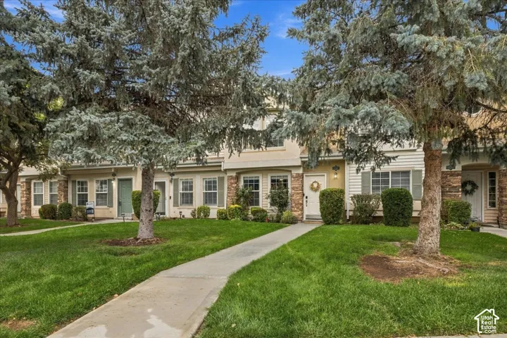 Manicured lawn with mature trees, concrete walkway, and townhome-style residences featuring light siding and stone accents