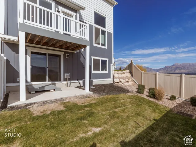 Two-tone exterior with white siding and dark gray panels, second-story balcony with white railing, ground-level patio with sliding glass door, fenced yard with landscape rock and manicured shrubs, mountain backdrop
