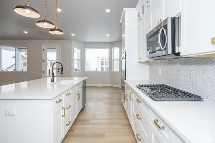 White kitchen cabinetry with gold-tone hardware, stainless steel appliances, herringbone tile backsplash, and light wood-finish flooring