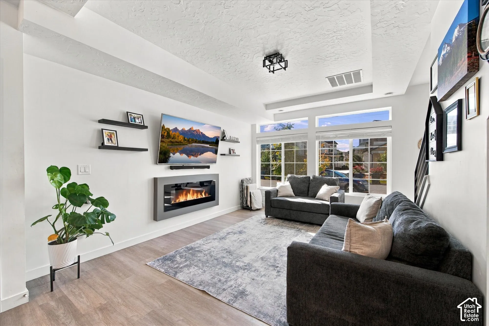 Recessed ceiling with integrated lighting, wood-finish flooring, large multi-pane windows, built-in electric fireplace, and dark-framed floating shelves