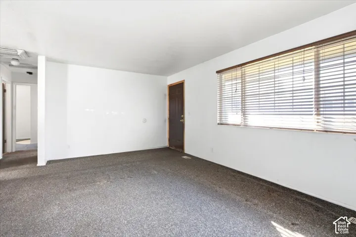 Spacious room featuring neutral white walls, dark-toned entry door, and a multi-pane window with horizontal blinds