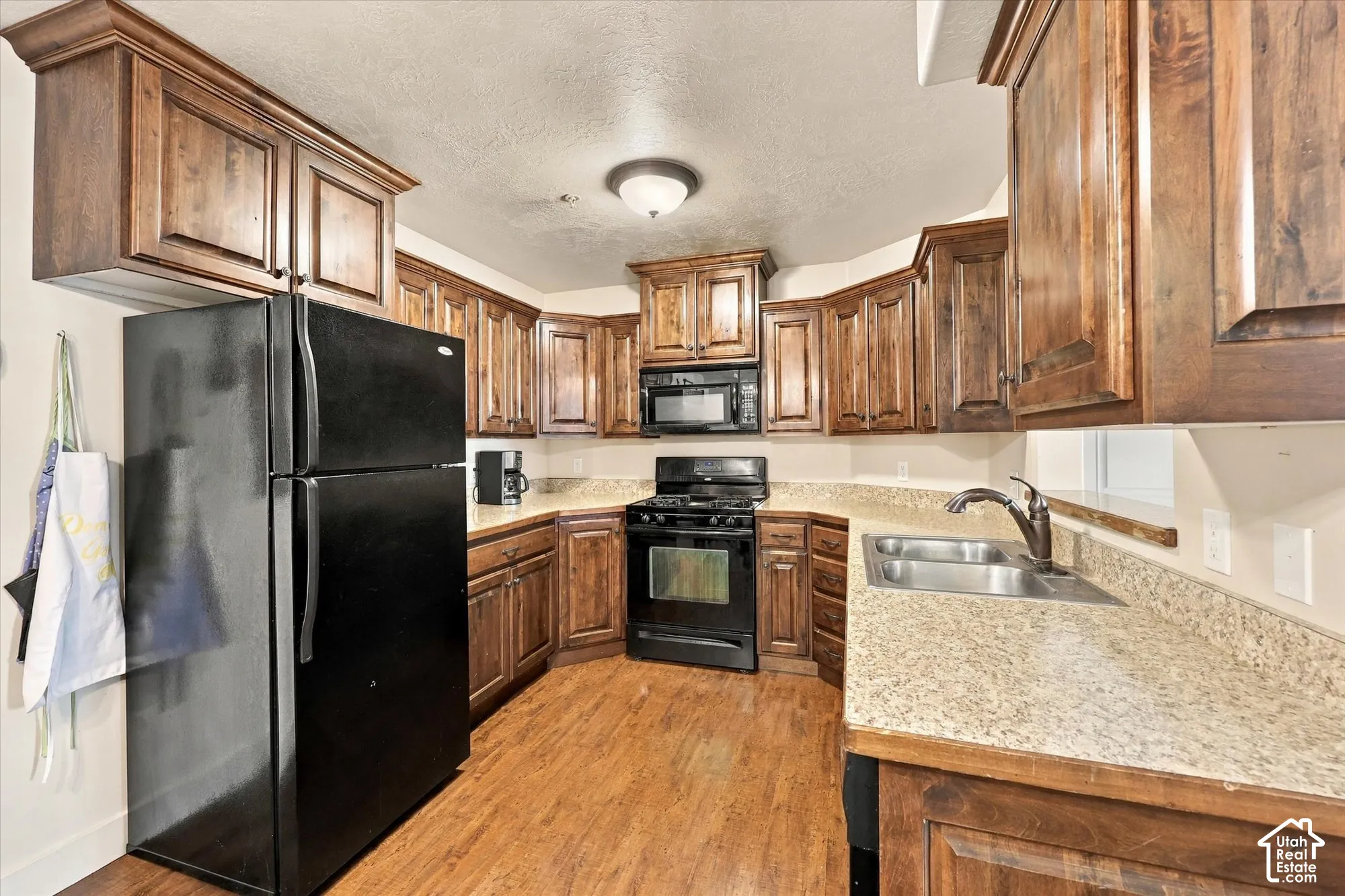 Kitchen featuring wood-finish cabinetry, granite-style countertops, black appliances, a stainless steel double basin sink, and wood-finish flooring