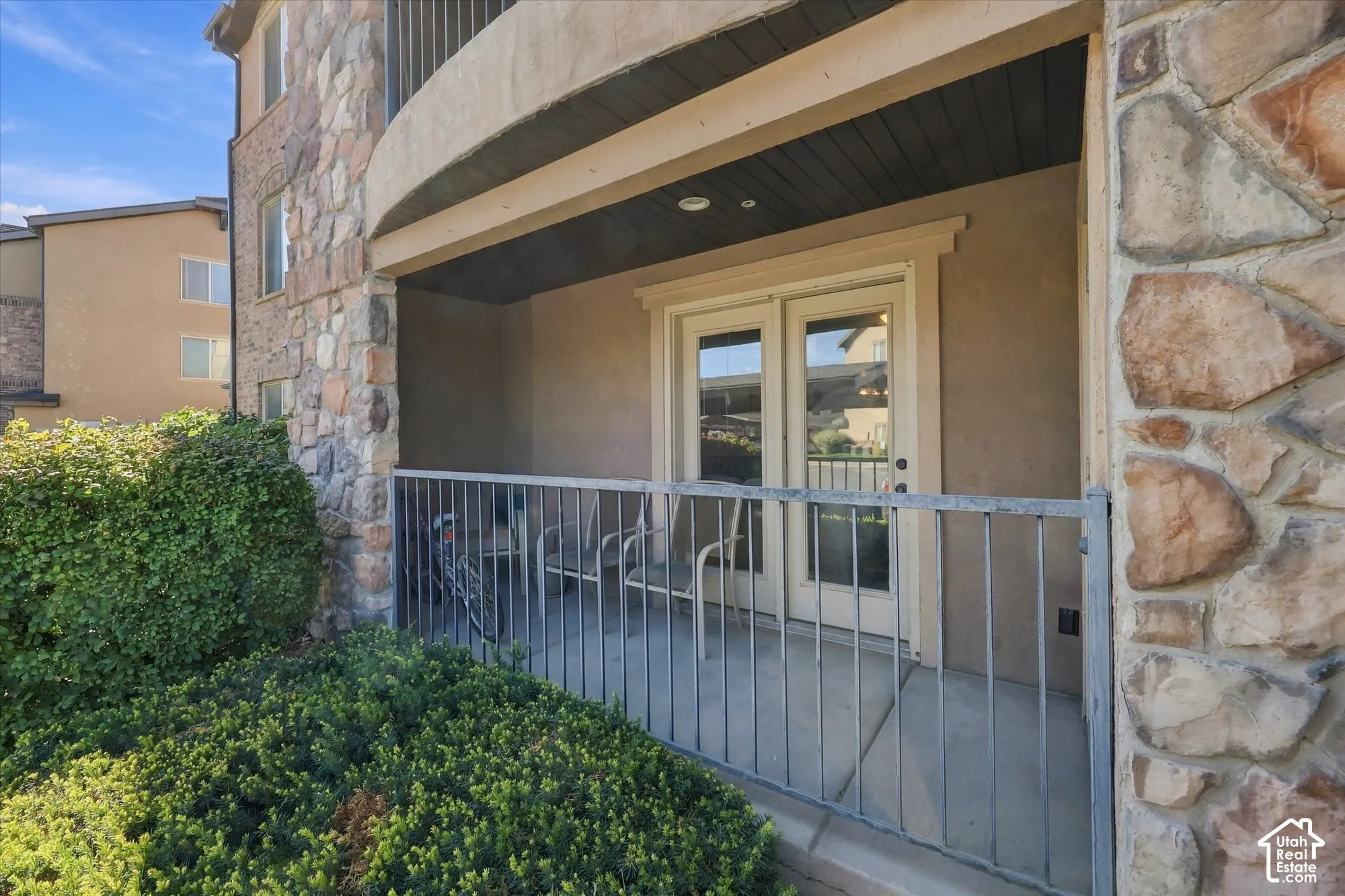 Private covered patio with concrete flooring, framed by mature shrubs and a stone-clad exterior
