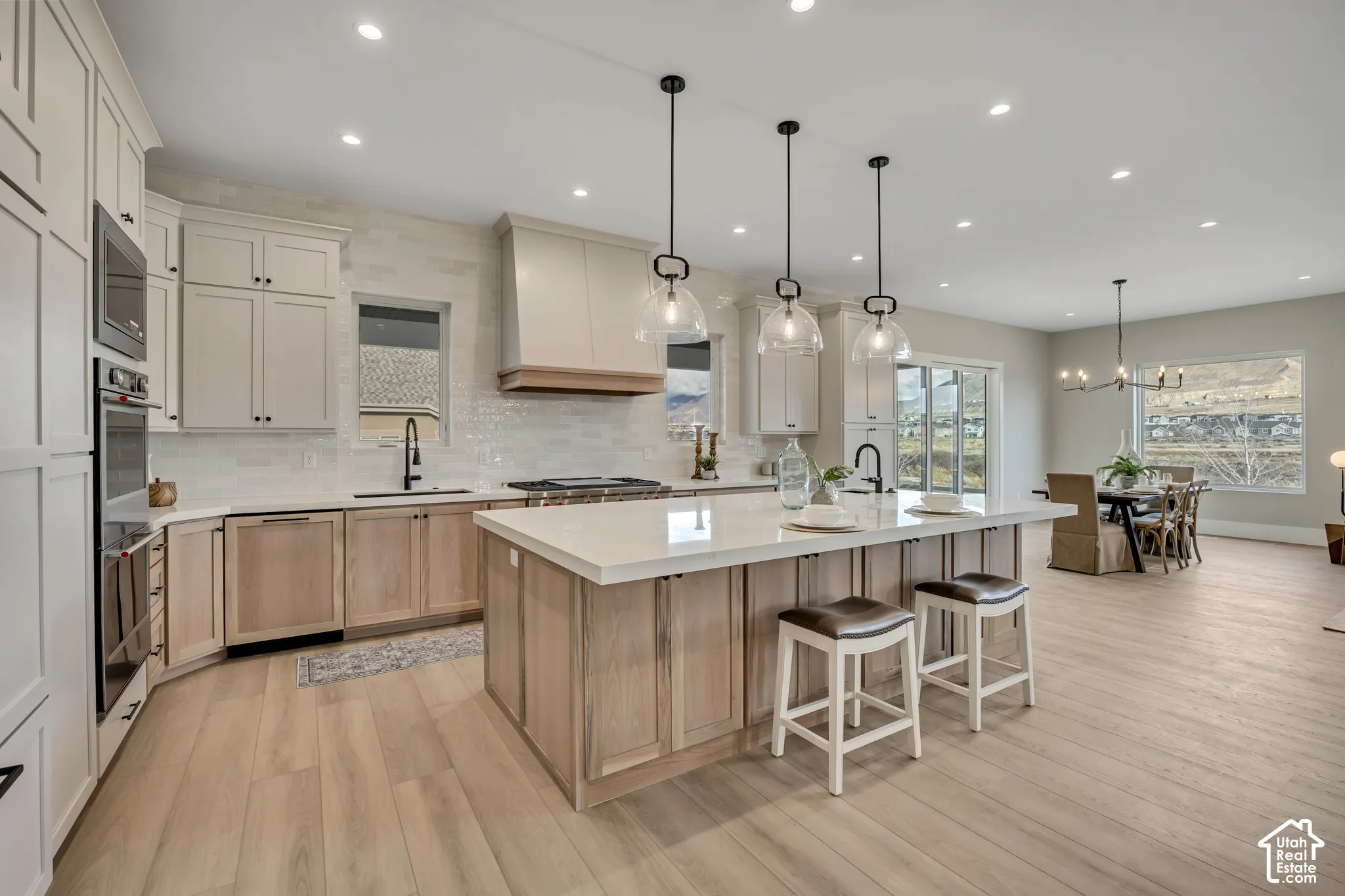 Expansive kitchen featuring light wood-finish flooring, a large center island with counter seating, light-toned cabinetry, stainless steel built-in appliances, and three pendant lights