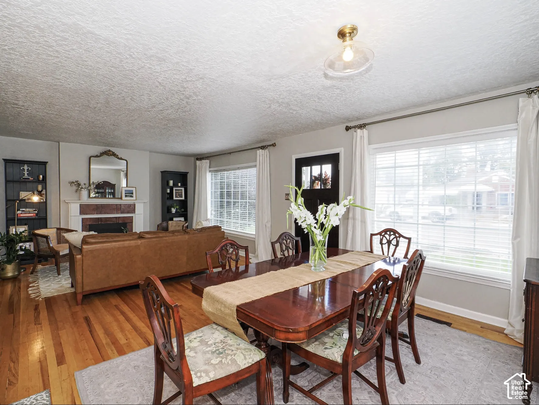 Hardwood flooring, fireplace with white mantel and brick surround, dark-paneled door, and two large windows with white blinds and drapes