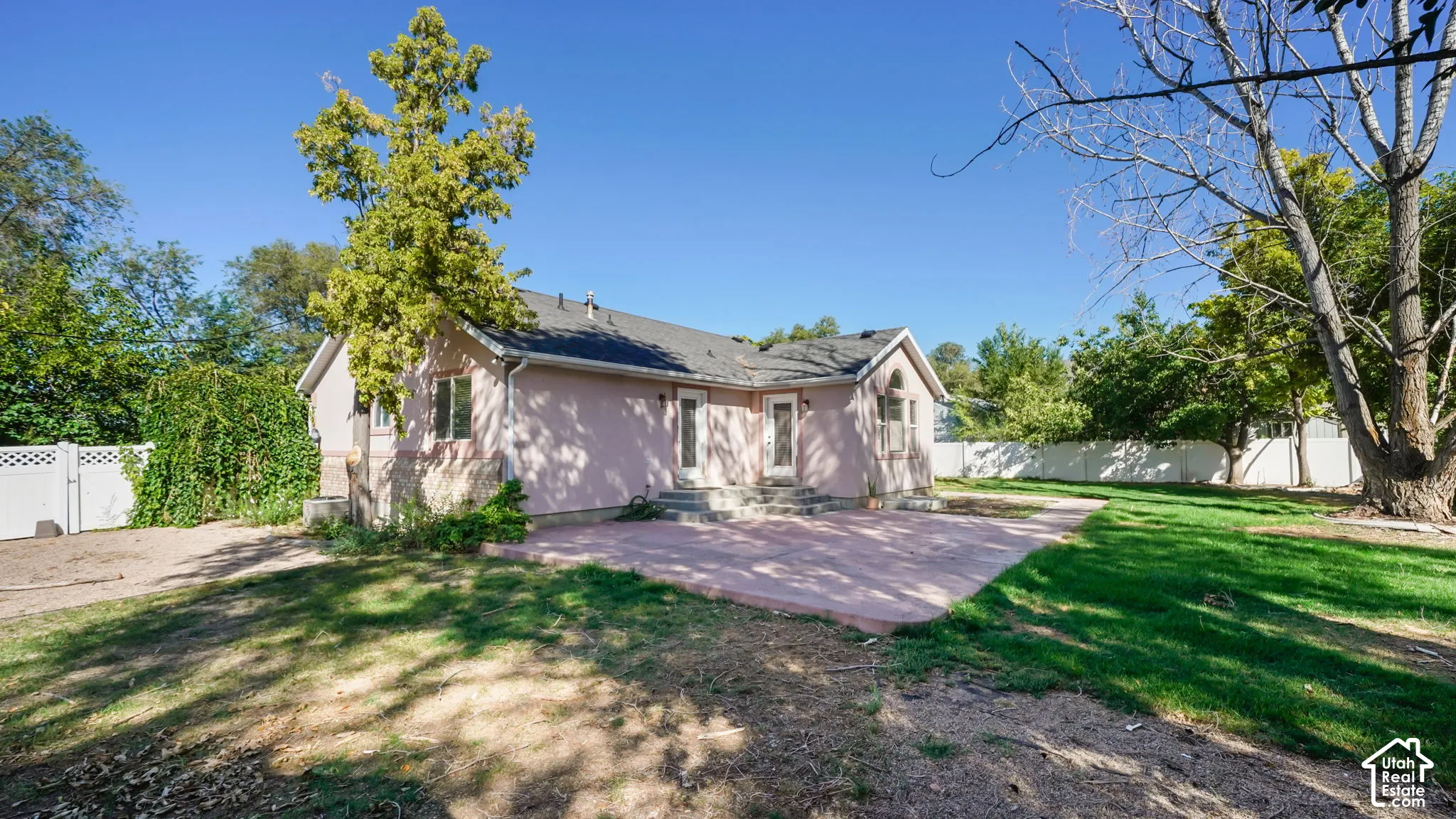 Expansive backyard featuring a large paved patio, mature trees, manicured lawn, and white privacy fencing