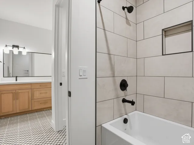 Bathtub with matte black fixtures, tiled surround with recessed niche, wood-finish vanity with countertop, and decorative floor tiling