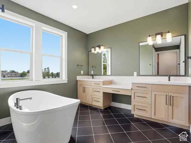 Freestanding soaking tub, dual windows, wood-finish vanity cabinetry, dual mirrors with integrated lighting, and dark tile flooring