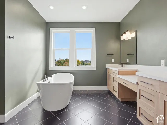 Freestanding soaking tub, large window, dual vanity with solid surface countertops, custom cabinetry, and tile flooring