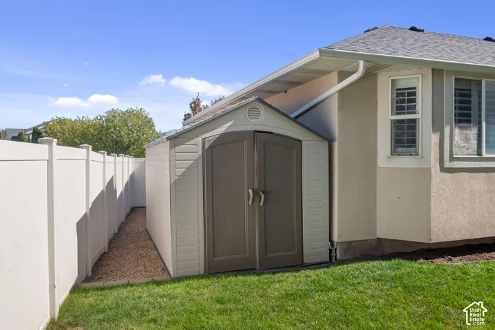 Detached storage shed with double doors, stucco exterior, white vinyl fencing, and a manicured lawn