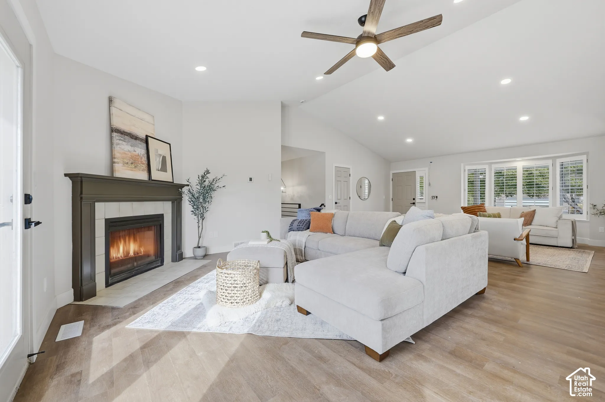Living area featuring a fireplace with a tiled surround, vaulted ceiling with a ceiling fan, recessed lighting, wood-finish flooring, and multiple windows with white shutters