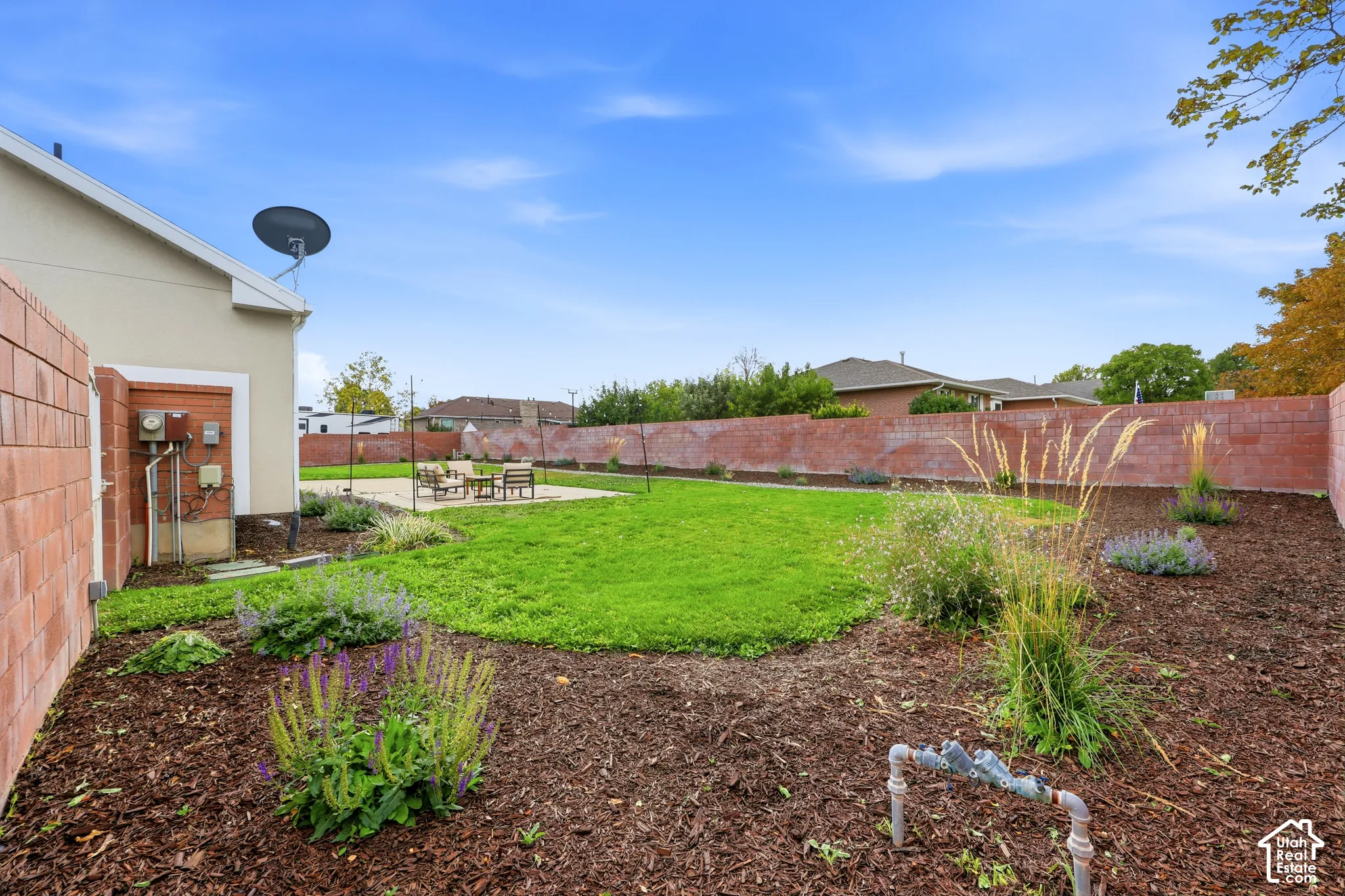 Expansive backyard featuring a manicured lawn, brick perimeter wall, concrete patio, and garden beds with mature plantings