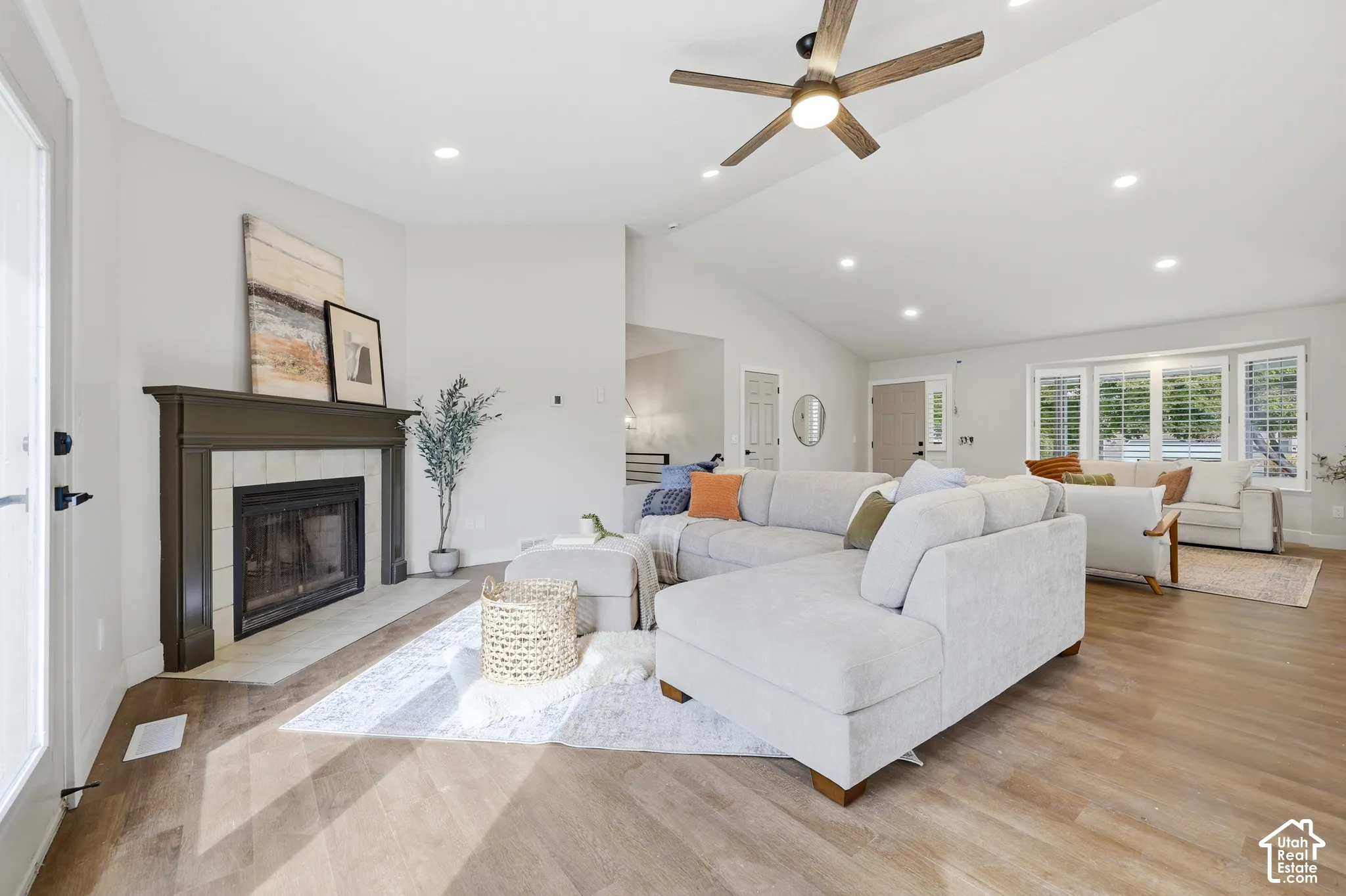 Vaulted ceiling with a ceiling fan, recessed lighting, wood-finish flooring, white tiled fireplace with a dark mantel, and multiple windows