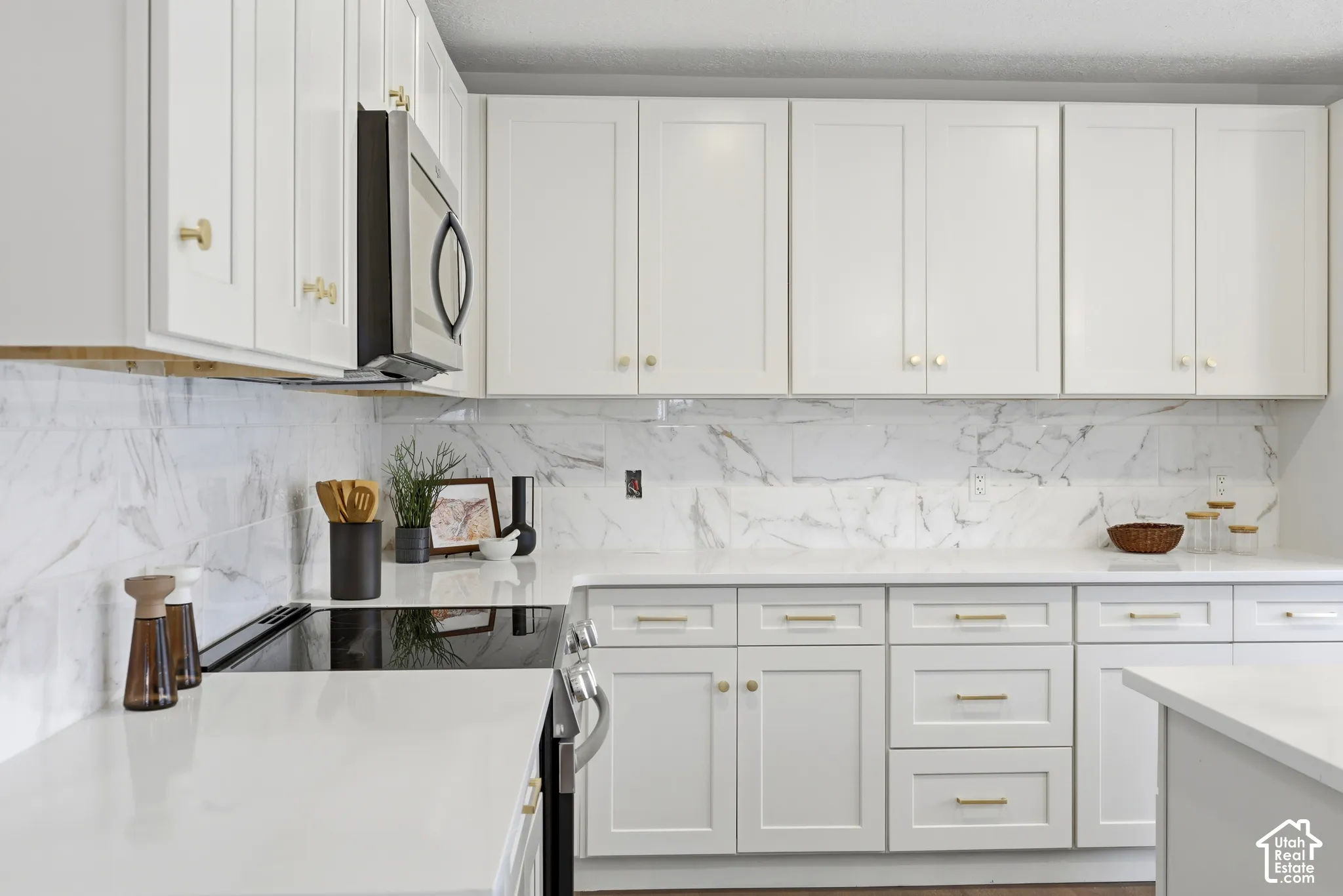 White shaker-style cabinetry with gold-tone hardware, white countertops, a large format marbled backsplash, and stainless steel appliances