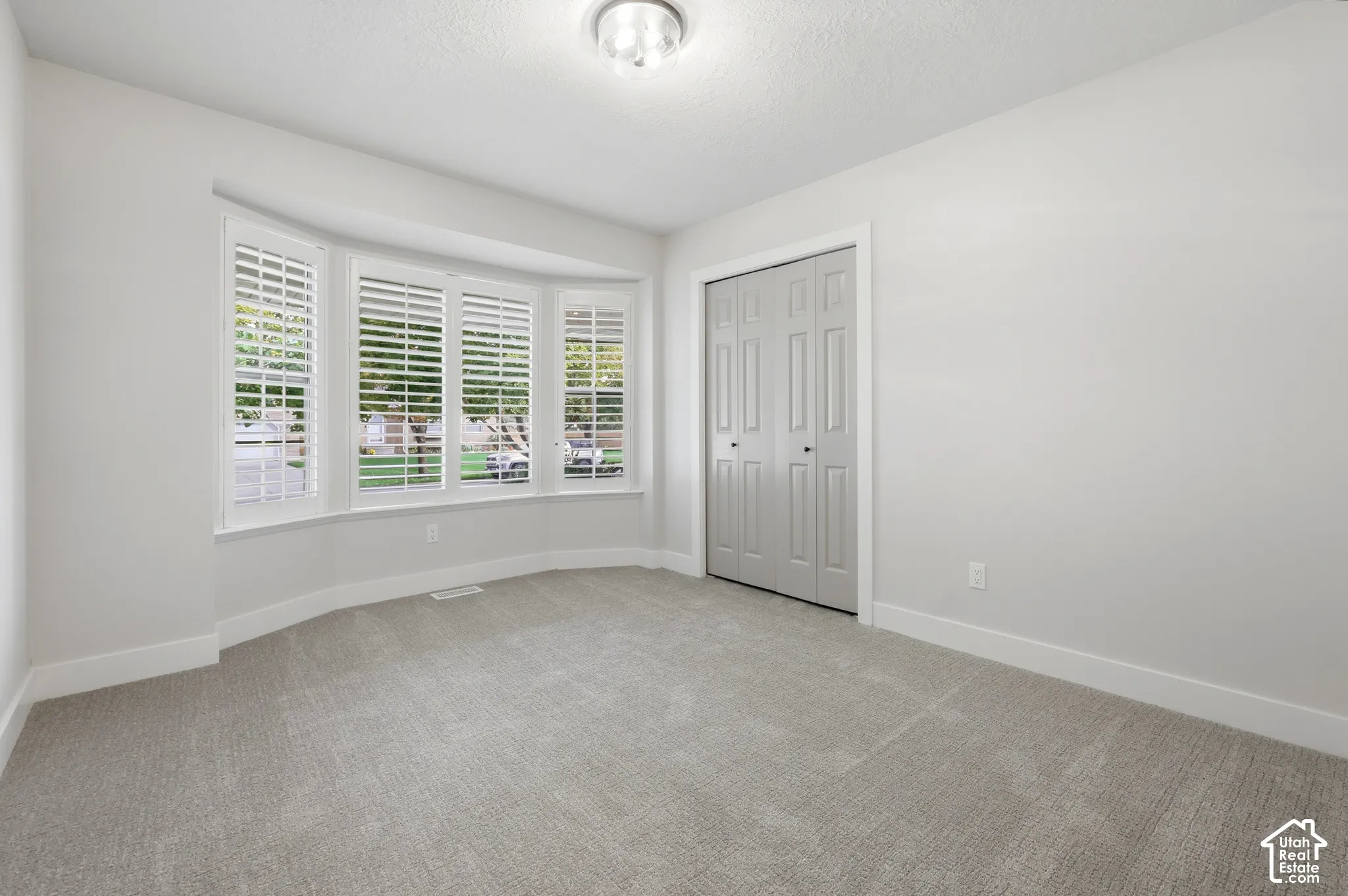 Recessed bay window with plantation shutters, neutral tone carpeting, white walls, bi-fold closet doors, and a flush mount light fixture