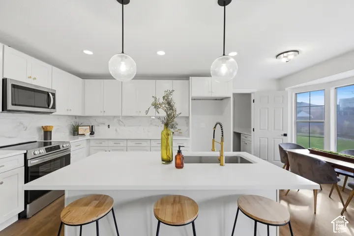 White shaker cabinetry with gold hardware, stainless steel appliances, a large center island with seating, a gold-finish gooseneck faucet, and wood-finish flooring