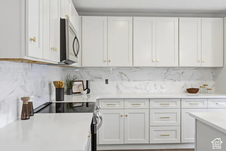 White shaker-style cabinetry with gold-tone hardware, white countertops, a large format marbled backsplash, and stainless steel appliances