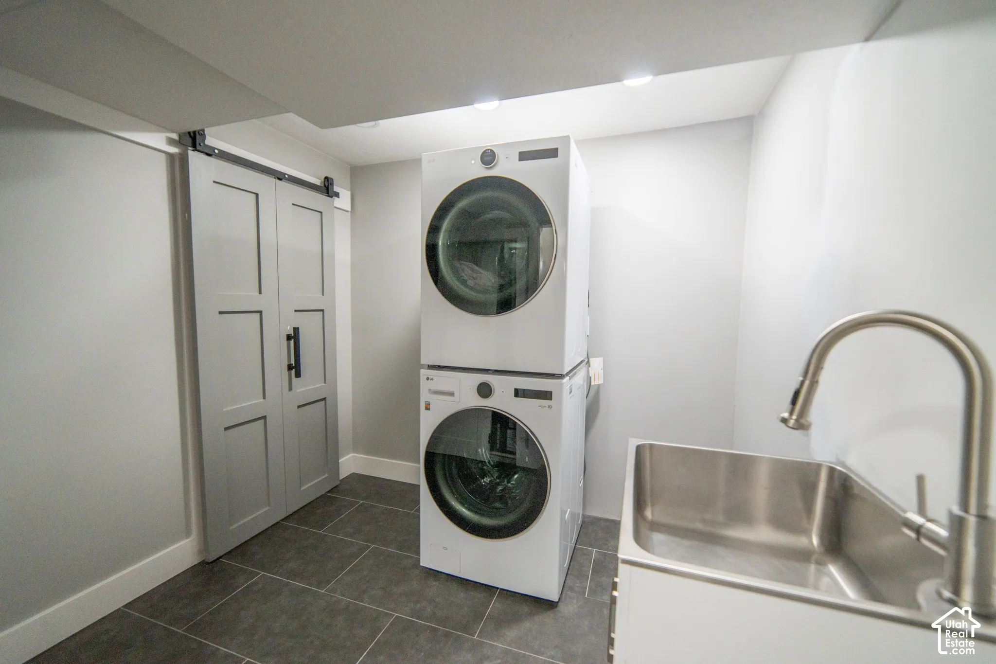 Barn door, stacked laundry appliances, stainless steel utility sink, pull-down faucet, and tiled flooring