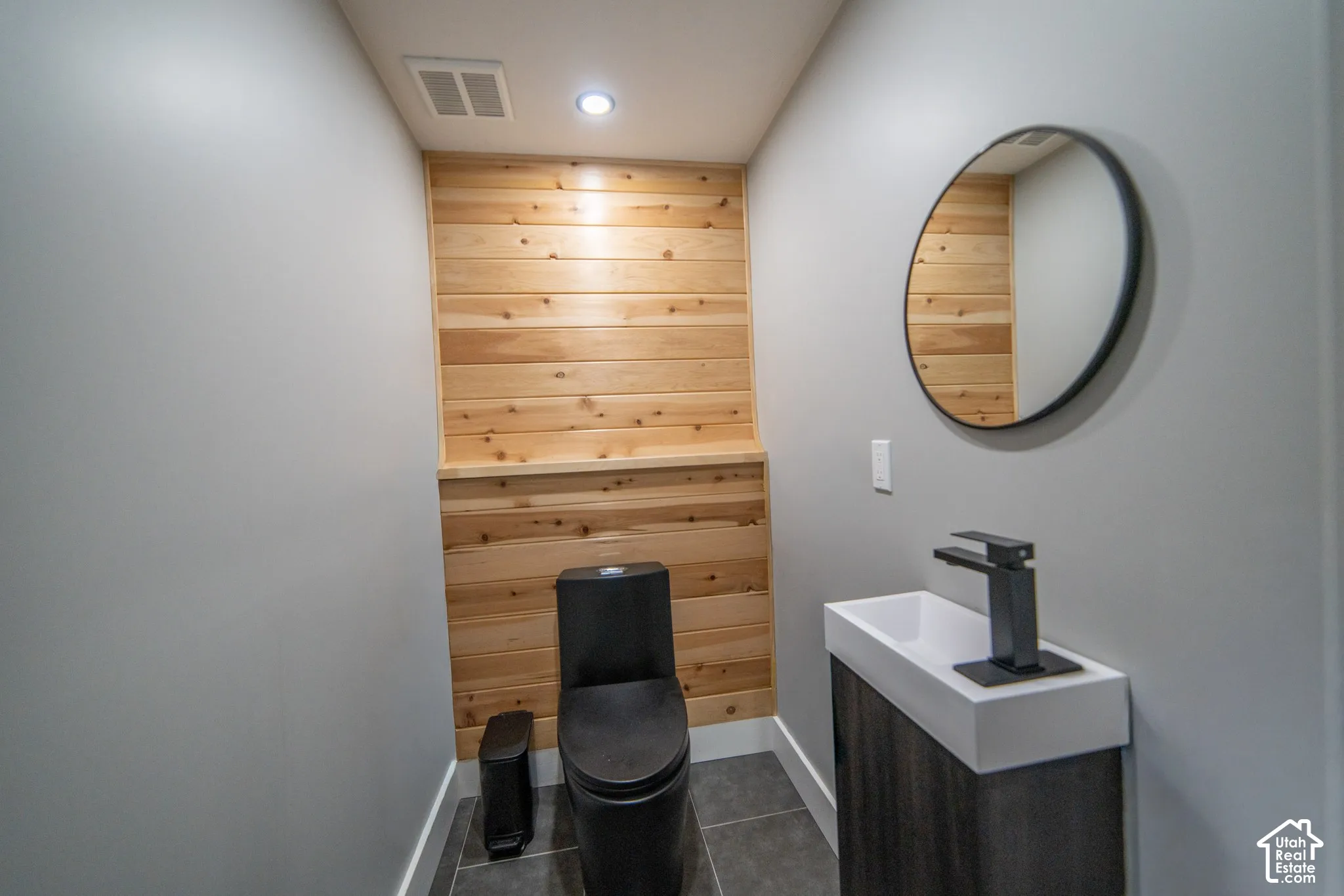Wood panel accent wall, dark gray floor tiling, circular wall mirror, wall-mounted sink with dark finish faucet, dark finish toilet