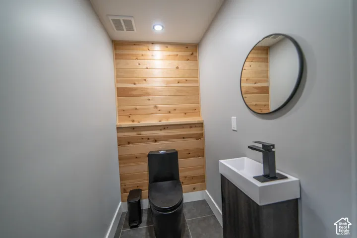 Wood panel accent wall, dark gray floor tiling, circular wall mirror, wall-mounted sink with dark finish faucet, dark finish toilet