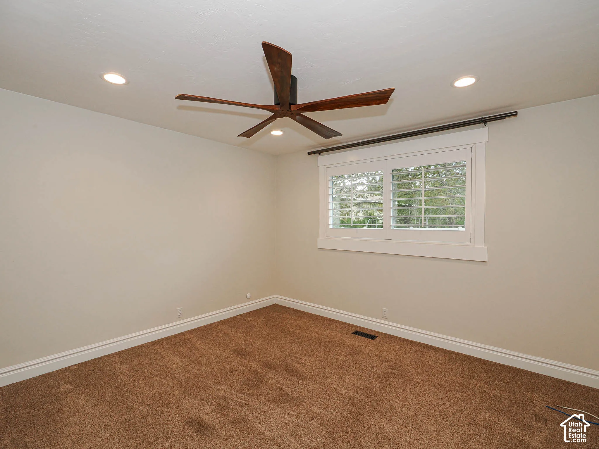 Recessed ceiling lighting, wood-finish ceiling fan, window with white plantation shutters, and brown carpet flooring
