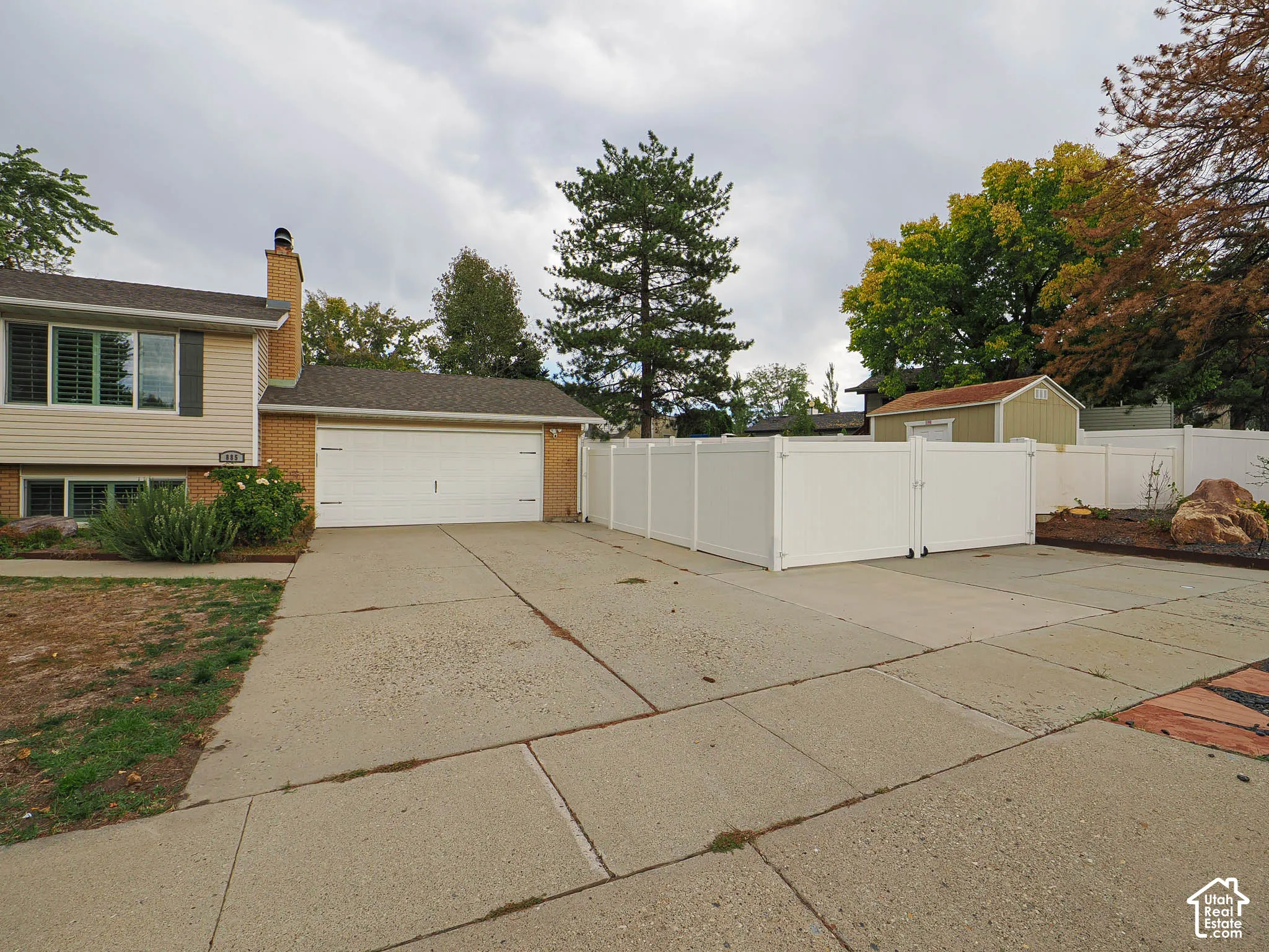 Attached two-car garage, brick and siding exterior, concrete driveway, white privacy fencing, and an exterior storage shed
