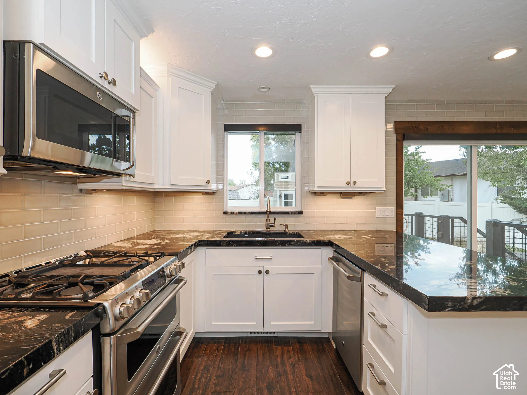 White cabinetry with dark contrasting countertops, stainless steel appliances, full tile backsplash, recessed ceiling lighting, and wood-finish flooring