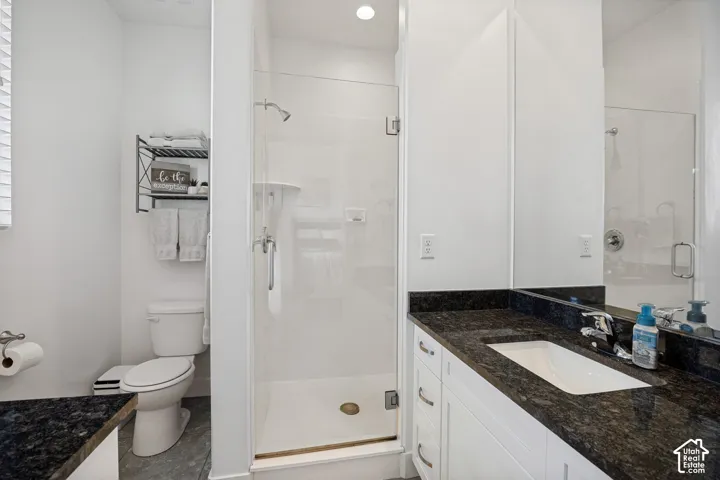 Glass-enclosed shower, white vanity with integrated sink, dark stone countertop, white toilet, and gray floor tiling