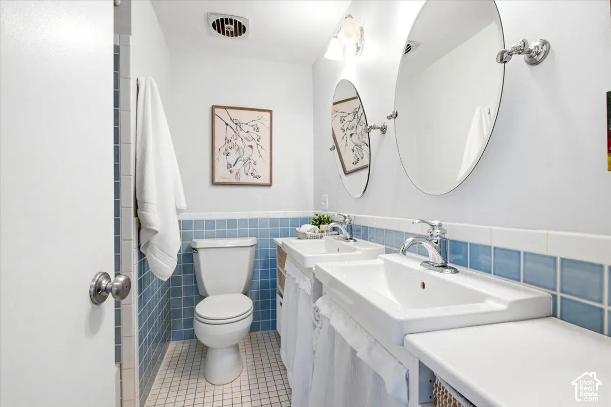Dual pedestal sinks with chrome fixtures, oval mirrors, white tile flooring, and blue square wall tiling