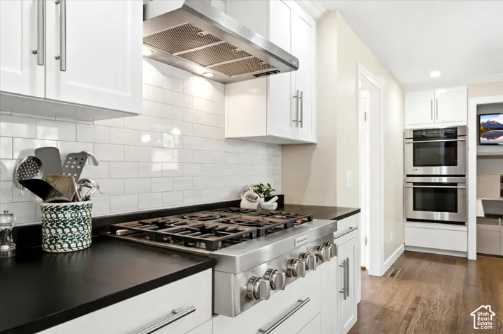 Stainless steel range with a ventilation hood, white shaker cabinetry, black countertops, white subway tile backsplash, and wood-finish flooring