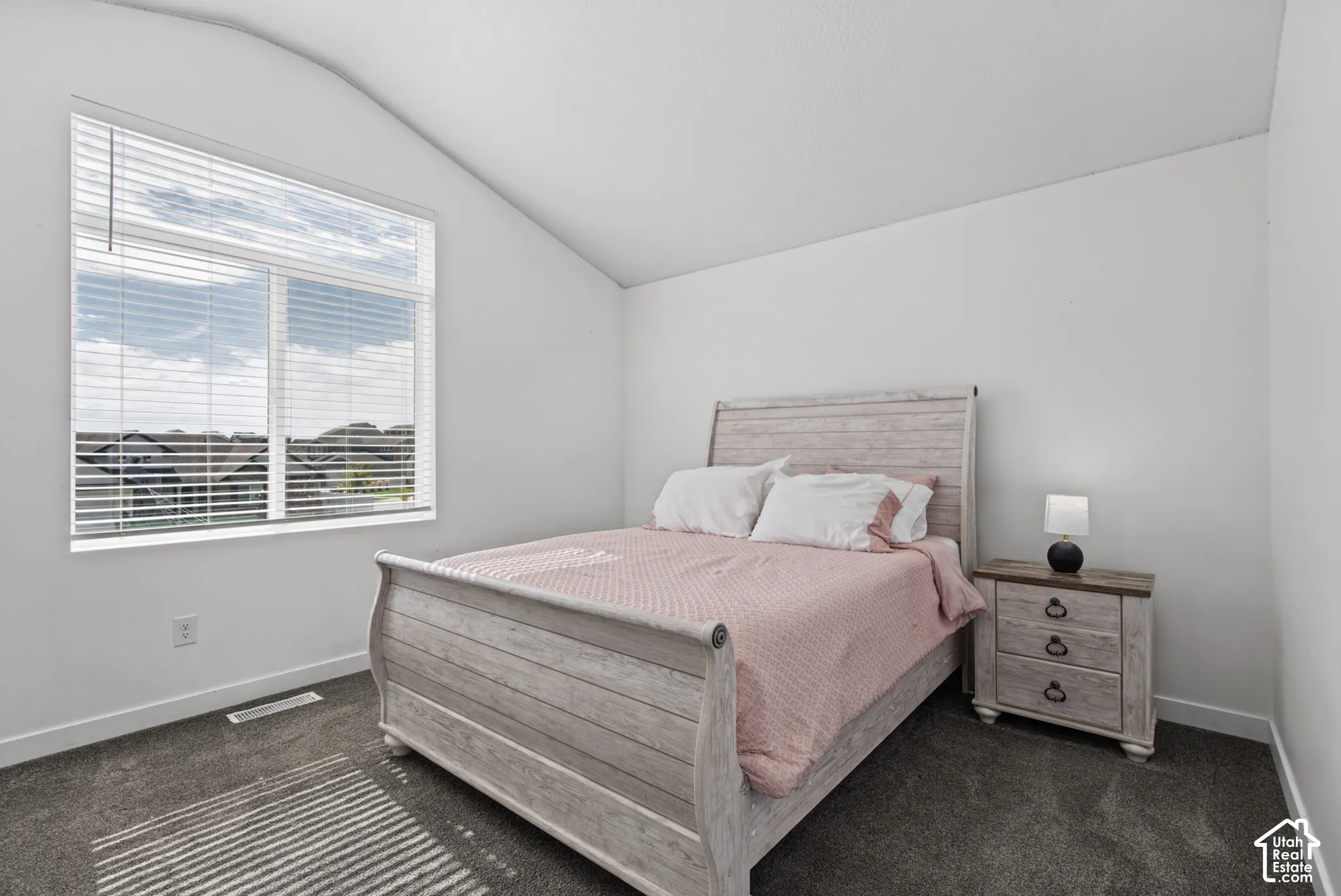 Angled ceiling, large window with blinds, light-colored walls, and dark carpet flooring