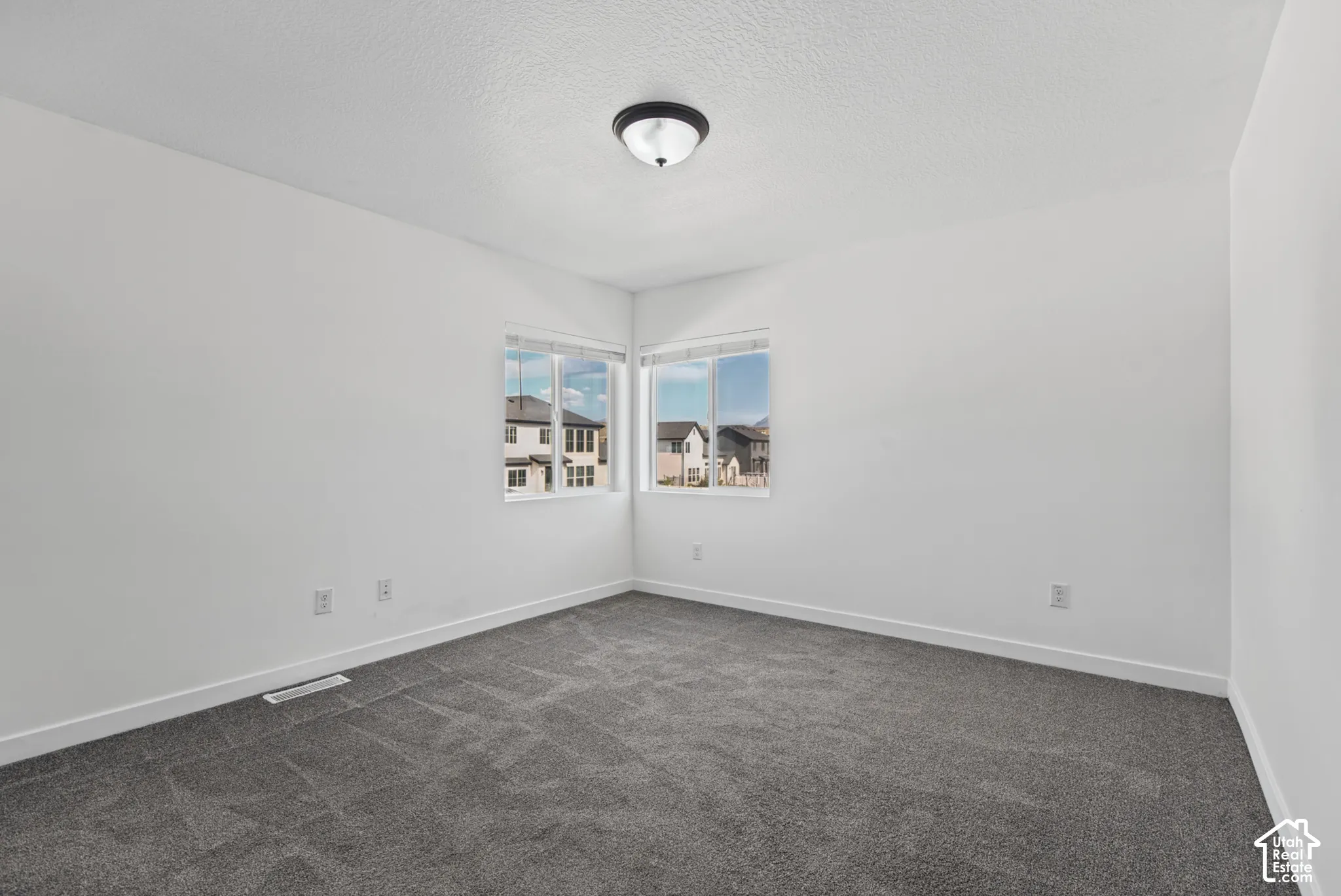 Neutral wall paint with textured finish, corner window placement, and dark gray carpet flooring