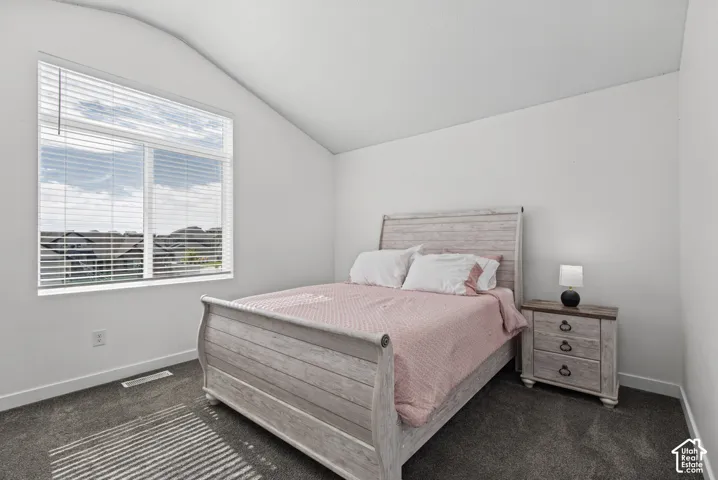 Angled ceiling, large window with blinds, light-colored walls, and dark carpet flooring