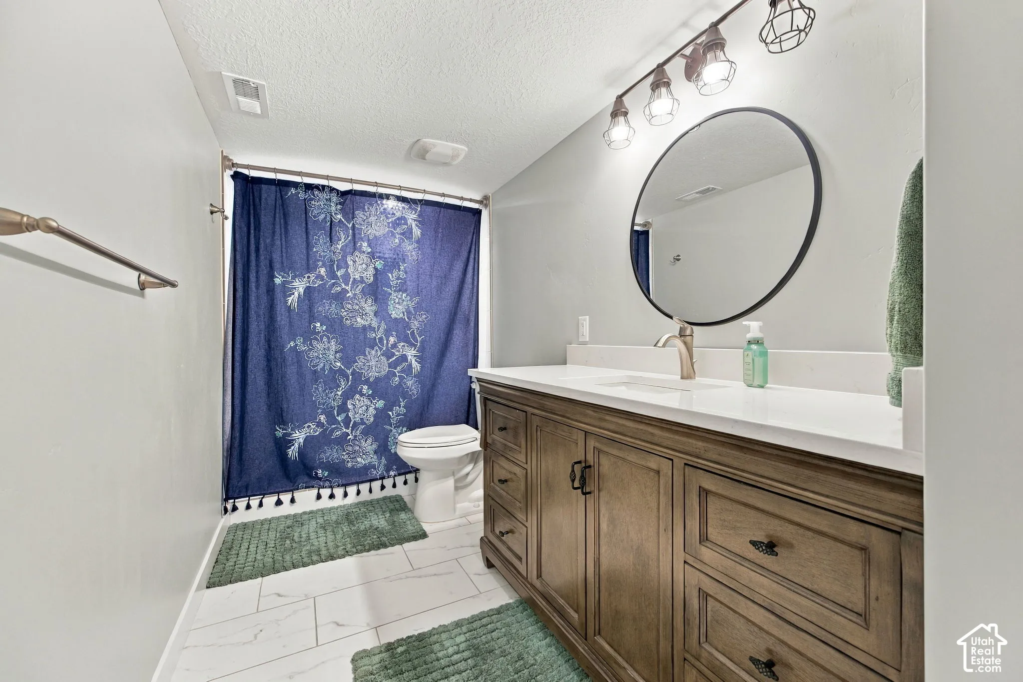 Wood-finish vanity with white countertop, round mirror, track lighting, and tile flooring