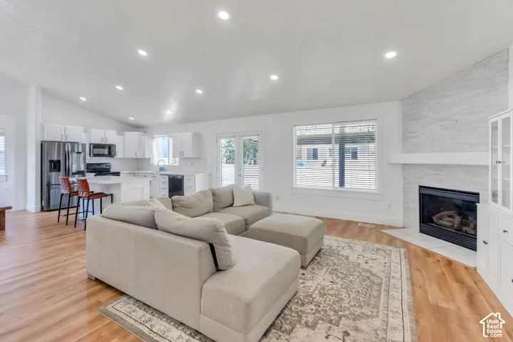 Open-concept living area with vaulted ceilings, recessed lighting, wood-finish flooring, a contemporary fireplace with a stone surround, and an adjacent kitchen featuring white cabinetry and stainless steel appliances