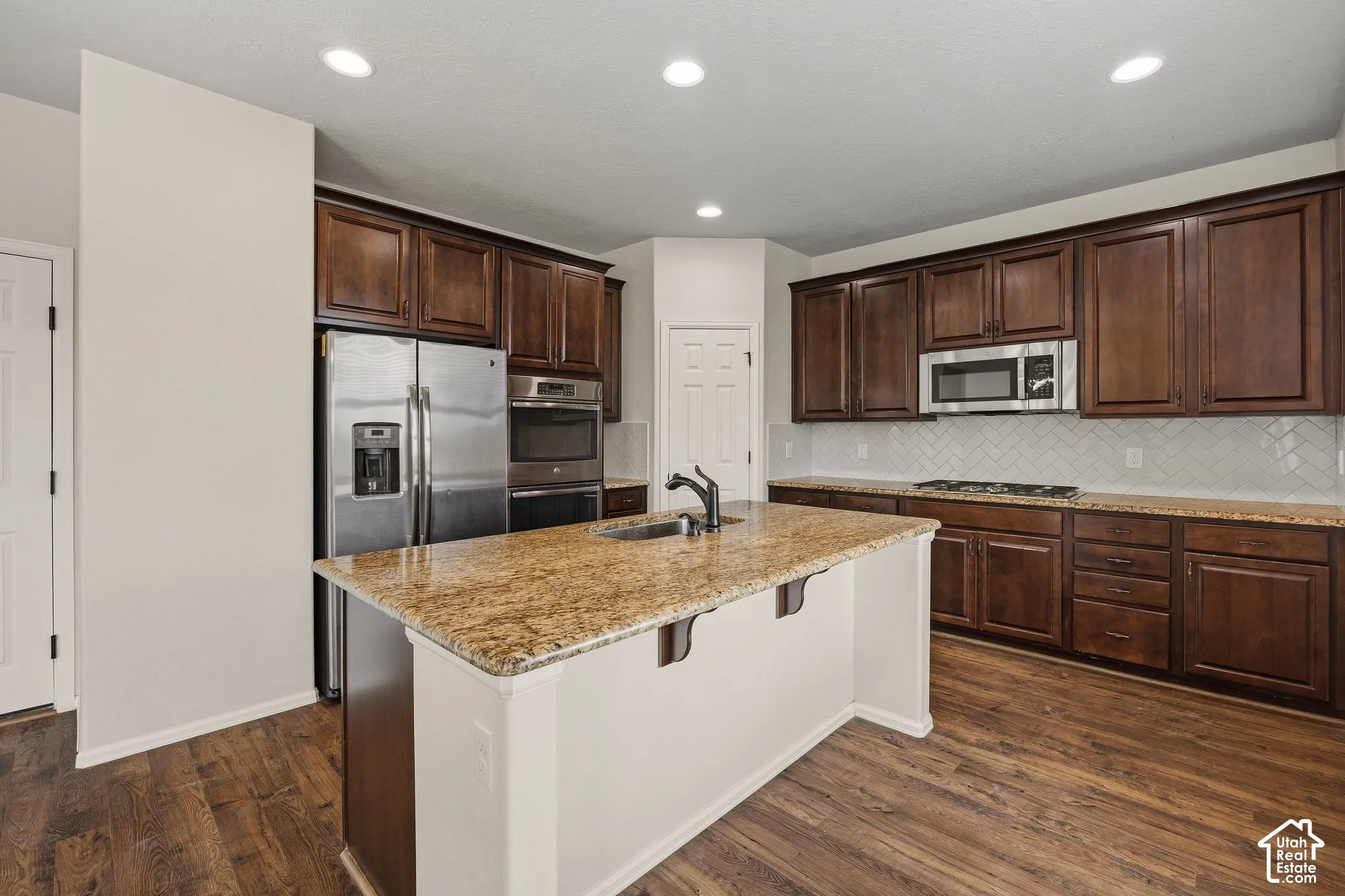 Kitchen featuring wood-finish flooring, dark wood cabinetry, stainless steel appliances, a center island with sink, and light-toned countertops