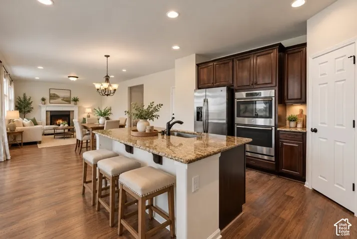 AI Staged Kitchen featuring a stone-topped island with an undermount sink, dark wood cabinetry, built-in stainless steel double ovens, family room with fireplace and wood-finish flooring