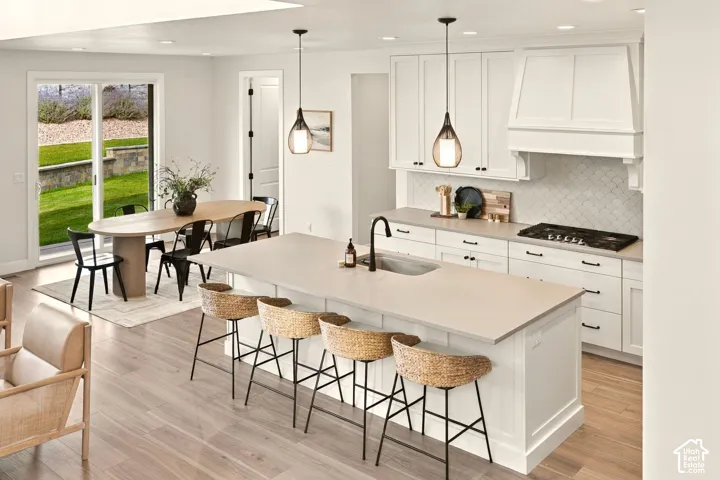 Open-concept kitchen and dining area featuring light wood-finish flooring, a kitchen island with an undermount sink, white shaker-style cabinetry, and a tile backsplash