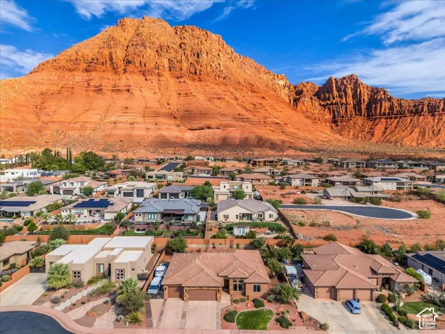 Desert landscape setting with a prominent red rock mountain backdrop, residential community with diverse architectural styles, multiple homes featuring solar panels, and integrated landscaping with desert plantings