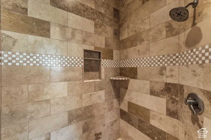 Spa-inspired shower featuring travertine tile walls in mixed earth tones, decorative mosaic tile accent band, recessed shelving niche, rainfall showerhead, and lever-handle valve trim in brushed bronze