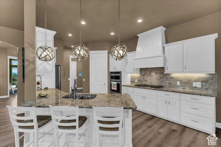 Kitchen featuring a large island with sink, white shaker cabinetry, stone countertops, stone tile backsplash, and wood-finish flooring