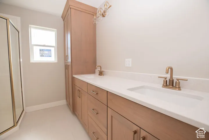 Dual vanity with integrated sinks and brushed brass fixtures, light-toned wood-finish cabinetry, built-in storage tower, and a clear glass shower enclosure